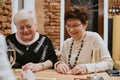 Two smiling elderly dark-haired and fair-haired women sit at wooden table with glasses of champagne playing dominoes.