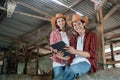 two smiling farmers in casual clothes sit by a wooden fence while using a digital tablet