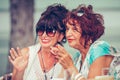 Two smiling female senior friends talking and pointing fingers to someone while drinking coffee Royalty Free Stock Photo