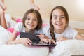 Portrait of two smiling girls lying on bed with digital tablet and looking in camera Royalty Free Stock Photo