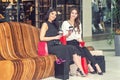 Two smiling girls sits on the wood bench with bags while looking at camera at shopping center Royalty Free Stock Photo