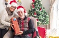 Smiling senior retired peoples with Santa`s cap sitting and showing the received gift. Gifts and Christmas treee in the backgroun