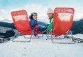 Two smiling skiers sit in chaise-longue on the mountain top