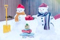 Two smiling snowman - happy couple  in knitted mittens, scarf and red caps with gifts and shovel  in the back yard Royalty Free Stock Photo