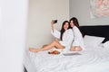 Two smiling women in white bathrobes happily taking photos together on cellphone with delicious pastry and coffee Royalty Free Stock Photo