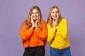 Two smiling young blonde twins sisters girls in vivid colorful clothes putting hands on cheeks isolated on pastel violet Royalty Free Stock Photo