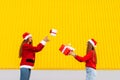 Two smiling young women in red sweaters and santa claus hats give each other christmas gifts while standing against yellow wall Royalty Free Stock Photo