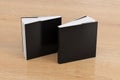 Two softcover or paperback square black mockup books standing on the wooden background. Blank front and back cover