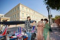 Two South Asian tourists standing with tablets happily negotiating fares with tuk-tuk drivers by the side of the road over the