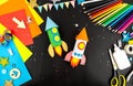 Two space rockets made of colored paper on a black table with colored pencils, colored paper, scissors and glue. The concept of