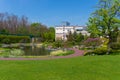 A two-storey white modern mansion near on a green lawn with a decorative pond and well-kept flower beds and trees under Royalty Free Stock Photo