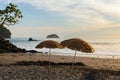 Two straw parasols planted on deserted beach seen at dusk, with small island in the Pacific Ocean in the background