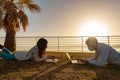 Two students lying on a meadow at sunset learning lesson using laptop computer in the city park of tropical ocean sea resort. Royalty Free Stock Photo