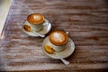 Two stylish white cups of cappuccino with latte art on saucer and golden spoons on light wooden table.