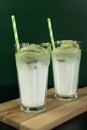 Two tall glasses with dalgona matcha and paper tubes on wooden board