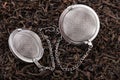 Two tea filter on a chain with black tea on a pile of dry tea leaves.