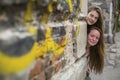 Two teen girls look out from behind the corner of a stone house.