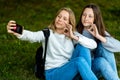 Two teenage girls. In summer in park nature. Sit on grass behind backpacks. In his hands holds smartphone. Take photos Royalty Free Stock Photo