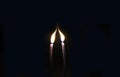 Two thin candles stretch their lights towards each other. The candles are in complete darkness.