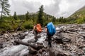 Two tourists with large backpacks cross the river in rubber boots. Royalty Free Stock Photo