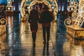 Two unrecognizable enamored young people going in neon lighting arch, city with festive illumination, evening. Christmas, holidays