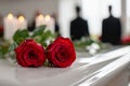 Two vibrant red roses resting on a white polished surface with blurred candles and somber figures in the background evoking a Royalty Free Stock Photo