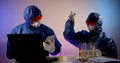 Two virologists are in the lab, sitting at a table with a laptop, notes, and flasks of analysis. They are wearing a blue Royalty Free Stock Photo