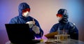 Two virologists are in the lab, sitting at a table with a laptop, notes and flasks of analysis. They are wearing a blue Royalty Free Stock Photo