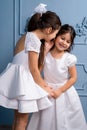 Two whispering kids in white dresses, on a gray background.