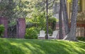 Two white adirondack chairs setting under tall trees on a shady hill in front of ornate iron garden fence and two story Tudor styl