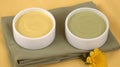 two white bowls filled with green and yellow liquid on top of a green napkin with a yellow flower on the side of the bowl and a