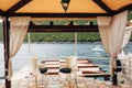 Two white chairs with black pillows stand in a gazebo by the sea overlooking a white boat, in Montenegro, on the Lustica