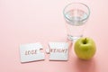 two white disconnected puzzles with the inscription lose weight, a glass of water and a green apple on a pink background