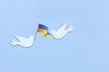 Two white figurines of a doves of peace with flag of Ukraine in their beaks on the light blue background. Peace Concept