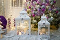 Two white lanterns standing at Christmas tree decorated with purple bulbs and warm garland lights. Blurry background with bokeh. Royalty Free Stock Photo