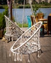 Two white macrame chair swings with tassels on bottom, and a garden furniture on a bright wooden patio, on the riverbank. Royalty Free Stock Photo