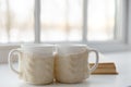 Two white mugs in a scarf stand on a table in the background of a window with cozy aroma of coffee.