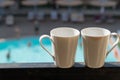 Two white mugs stand on the balcony in the background of the hotel, where the buildings and the pool are visible. On one mug there