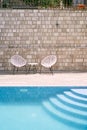 Two wicker chairs with a table stand near the stone wall by the pool