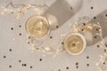 Two wineglasses with sparkling wine alcohol drink on neutral beige table with blurred garland lights, gold confetti
