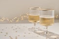 Two wineglasses with sparkling wine alcohol drink on neutral beige table with gold glitter confetti, blurred garland