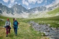 Two woman hikers are walking with trakking poles by the river among mountains. Extreme tourists in wild nature