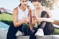 Two women athletes in sportswear sitting in park, relax after sports training, use smartphone, listening to music. Royalty Free Stock Photo