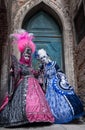 Two women in brightly colored masks and costumes standing in front of an old blue door in Venice during the Carnival