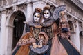 Masked women in costume, with decorated fans, standing in front of the arches at St Marks Square during the Venice Carnival