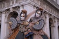 Two masked ladies in costume, with decorated fans, standing in front of the arches at St Marks Square during the Venice Carnival