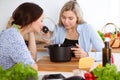 Two women friends looking into the dark pot with a ready meal and taste new recipes while sitting at the kitchen table Royalty Free Stock Photo