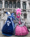 Two lovely women holding fans and wearing hand painted masks and ornate blue and pink costumes at Venice Carnival