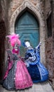 Two women in brightly colored masks and blue and pink costumes standing in front of an old blue door in Venice during the Carnival