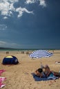 Two women people sitting under a blue and white beach umbrella against a bright blue sky